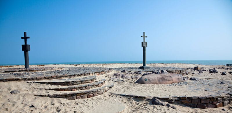 Two slender stone crosses on a paved area in the sand with the sea behind