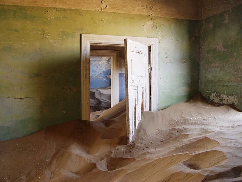 Verlassenes Zimmer mit abblätternder grüner Farbe, durch die offene Tür ist Sand weit in den Raum geweht